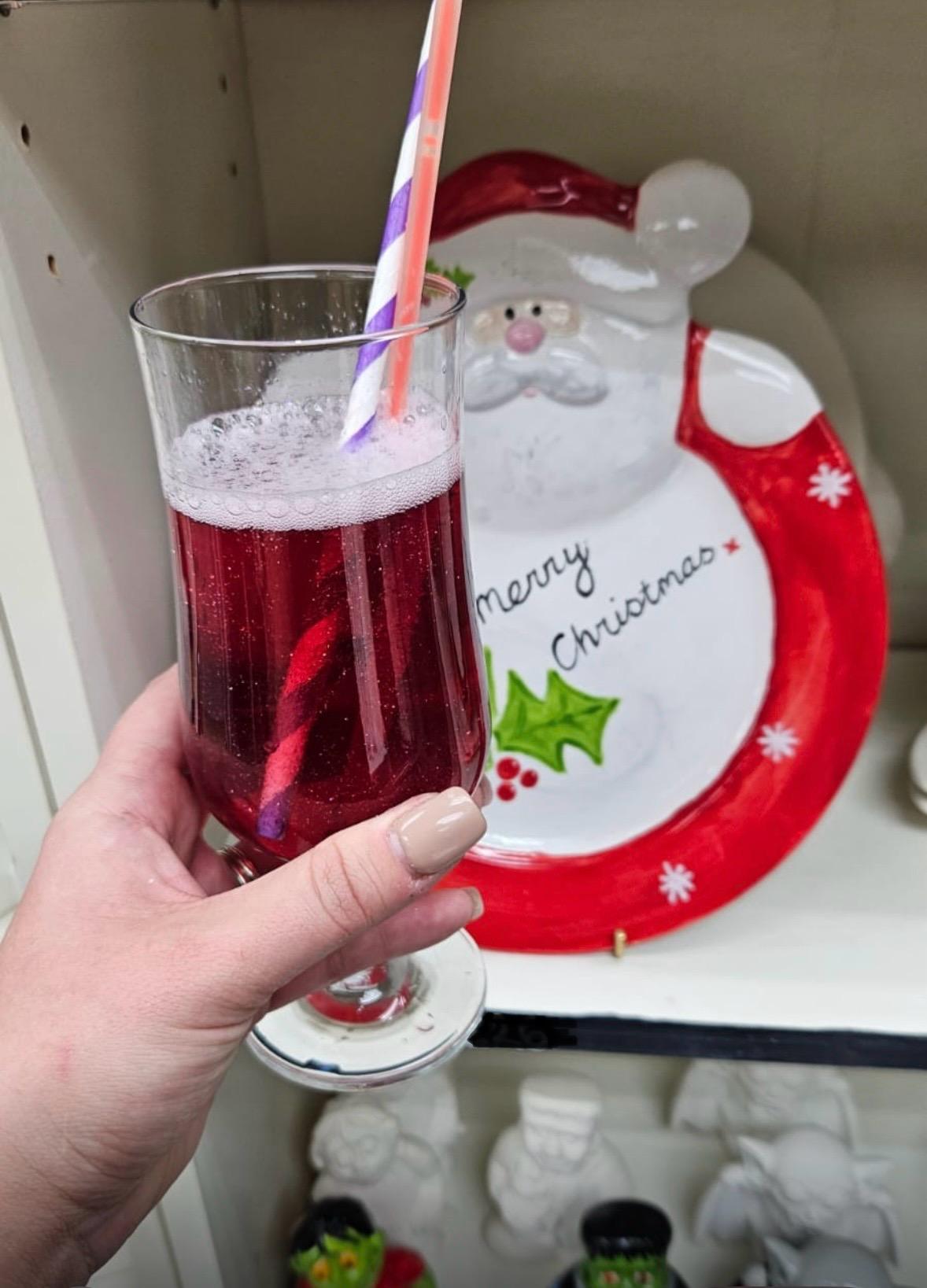 A red festive cocktail with straw in front of a Santa plate painted with Merry Christmas written on it